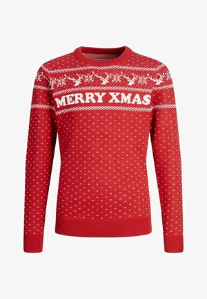 Osoba ubrana w czerwony sweter bożonarodzeniowy z napisem "MERRY XMAS", czarne spodnie dresowe oraz czarne wysokie trampki, stojąca z rękami w kieszeniach.