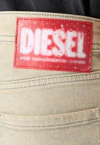 Tejido de mezclilla beige con una etiqueta rectangular roja que dice "DIESEL" en texto blanco, mostrando una superficie texturizada y gotas de agua.