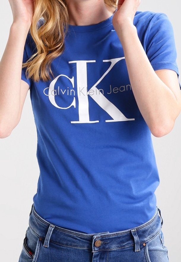 Person som bär en blå Calvin Klein Jeans t-shirt och blå jeans, justerar håret med båda händerna nära huvudet.