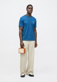 Modré bavlněné tričko s krátkými rukávy a vyšívaným logem; doplněné světlými béžovými kalhotami. Jedinec drží malý hnědý obal.