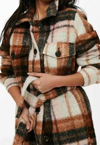 Manteau à carreaux marron et crème en tissu doux et texturé. Comprend de gros boutons, deux poches avant et une ceinture pour la fermeture.