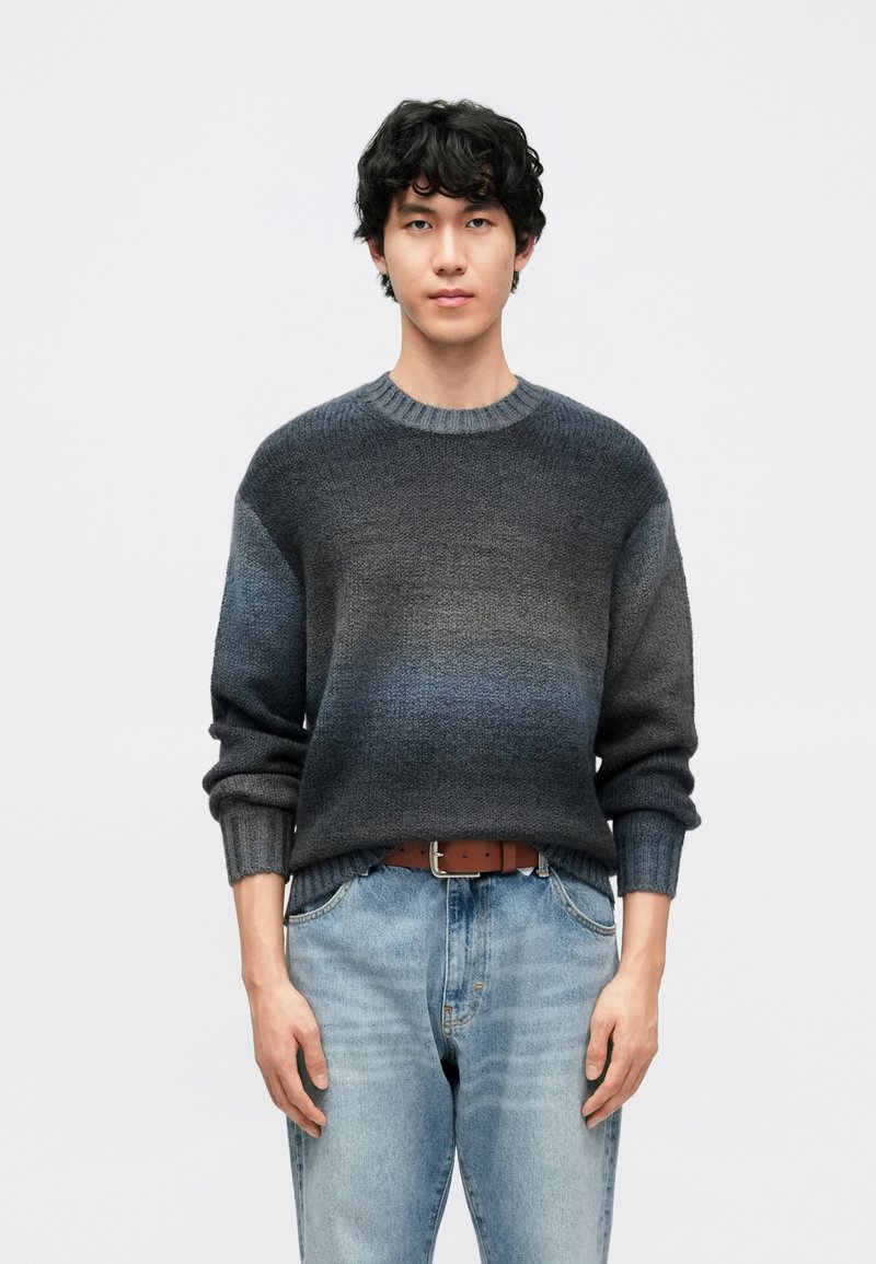 Temno sivi pulover z degradiranim učinkom, z rebrastim izrezom in manšetami, nošen čez svetlo modre jeans hlače. Tkanina je videti strukturirana in mehka.