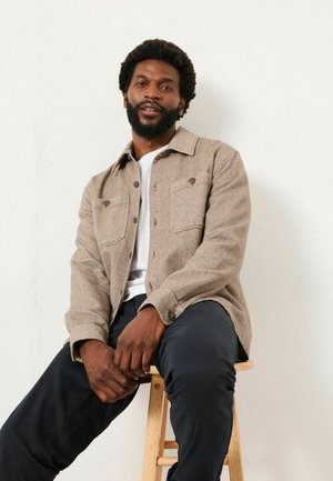 Man with beard wearing beige jacket, white shirt, and black pants sitting on wooden stool against plain wall.