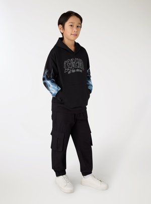 Bambino in piedi con le mani in tasca che indossa una felpa nera con cappuccio con la scritta "Legend of the Streets" e maniche con fulmini, pantaloni cargo neri, sneakers bianche.