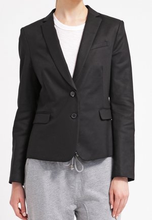 Person iført en tætsiddende sort blazer over en hvid skjorte kombineret med grå joggingbukser mod en ensfarvet hvid baggrund.
