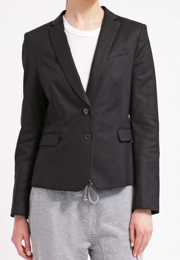 Personne portant un blazer noir ajusté sur une chemise blanche, associé à un pantalon de jogging gris, sur un fond blanc uni.