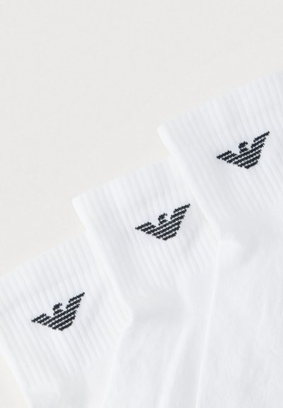 Calcetines de algodón blanco con textura acanalada, que presentan un diseño de logo con una raya negra en el puño. Tres pares dispuestos uno al lado del otro.