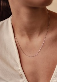 Collar de plata con un diseño liso y esbelto, reposando sobre la piel desnuda, combinado con una blusa de color claro texturizada que presenta un escote en V.