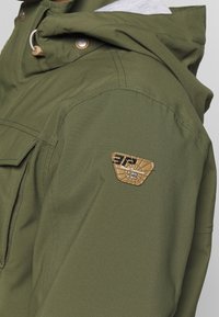 Épaule de veste vert olive avec un patch logo triangulaire marron et une partie d'une capuche avec un cordon et un bouton-pression visible.