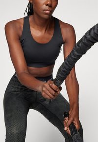 Sutiã desportivo preto com decote redondo, combinado com leggings pretas texturizadas apresentando um padrão de favo de mel subtil. O utilizador segura uma corda grossa.