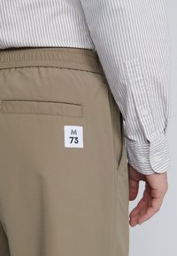 Beige bikses ar elastīgu jostu, ar aizmugures kabatu un baltu birku ar uzrakstu "M 73". Krekls ir gaiši pelēks ar plāniem svītrām.
