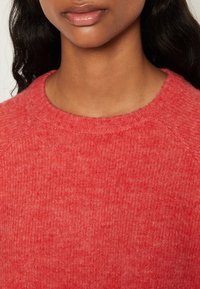 Punainen neulepaita, jossa on pyöreä pääntie ja pehmeä, teksturoitu viimeistely. Kangas on hieman meleerattu ja istuu rennosti.