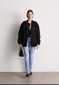 Μαύρο μπουφάν, ανοιχτά μπλε τζιν, μαύρα παπούτσια, κρατώντας μια μικρή μαύρη τσάντα. Το μοντέλο στέκεται μπροστά σε ένα ουδέτερο φόντο.
