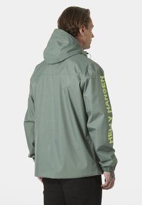 Chaqueta impermeable verde con capucha, que cuenta con puños elásticos y un brillante logo amarillo de "Helly Hansen" en la manga izquierda.