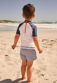 Kinder-Schwimmshirt: weiß mit marineblauen Ärmeln und orangefarbenem Reißverschluss; kombiniert mit blau-weiß gestreiften Schwimmshorts; barfuß am sandigen Strand.