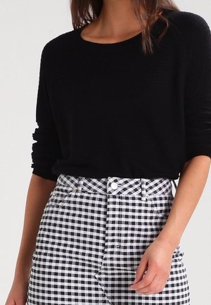 Frau trägt ein schwarzes Langarmshirt, das in einen hoch taillierten schwarz-weiß karierten Rock mit Knopfverschluss gesteckt ist.