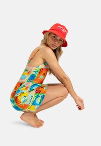Colorful dress with tropical postcard prints in orange, green, and blue; adjustable straps; worn with a red bucket hat featuring white text.