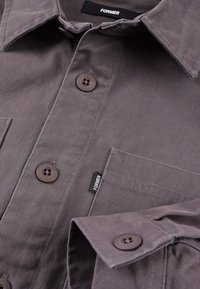 Γκρι πουκάμισο με κουμπιά, φτιαγμένο από υφασμάτινη υφή. Διαθέτει μυτερό γιακά, τσέπη στο στήθος και καφέ κουμπιά. Φέρει ετικέτα με την ένδειξη «FORMER».