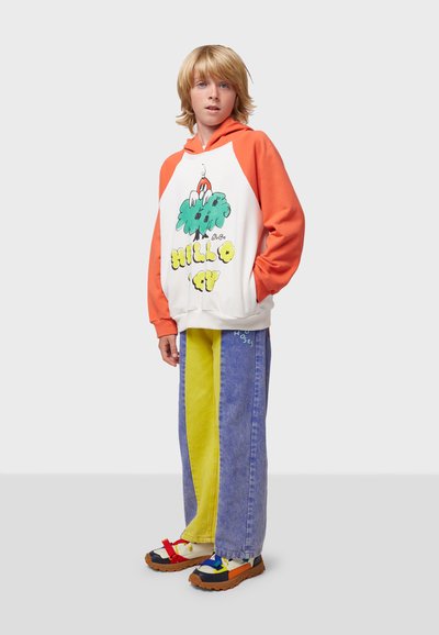 Sudaderacolorida con mangas naranjas y cuerpo blanco que presenta un gráfico divertido. Combinada con unos jeans bicolor coloridos y zapatillas multicolores.