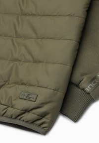 Giacca puffer trapuntata di colore verde oliva con una texture liscia. Presenta polsini a coste e un etichetta vicino all'orlo. Accenti in tonalità abbinate.