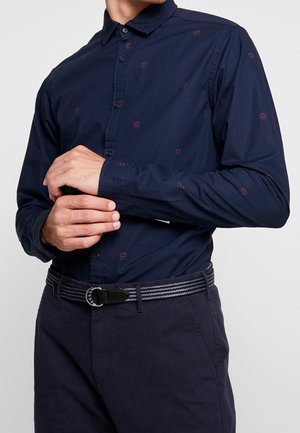 Homme ajustant le poignet d'une chemise à boutons bleu marine foncé avec de petits logos brodés rouges, portant un pantalon assorti bleu marine et une ceinture rayée.