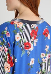 Donna con capelli rossi che indossa un abito floreale blu con fiori rossi, rosa e bianchi, accompagnati da foglie verdi e dettagli a strisce bianche e nere.