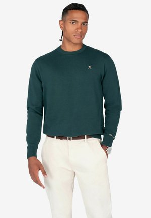 Suéter de cuello redondo verde oscuro con mangas largas, puños de canalé y un pequeño emblema en el pecho, combinado con pantalones de color claro.