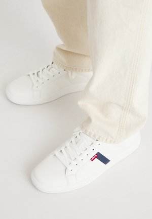 Persona che indossa sneakers bianche Levi's con lacci e pantaloni beige, in piedi su un pavimento bianco.