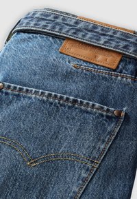 Denim jeans with a light blue wash, featuring a leather patch, golden stitching, and a curved back pocket design.