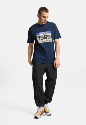 Młody mężczyzna patrzy w dół, ubrany w granatowy T-shirt z nadrukiem "1999", czarne spodnie dresowe i białe trampki z tęczowymi paskami.