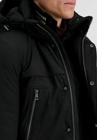 Zwarte gewatteerde jas met een hoge kraag, ritssluiting en een nepbontvoering. Heeft voorzakken en metalen ritsaccenten. Glad textuur.