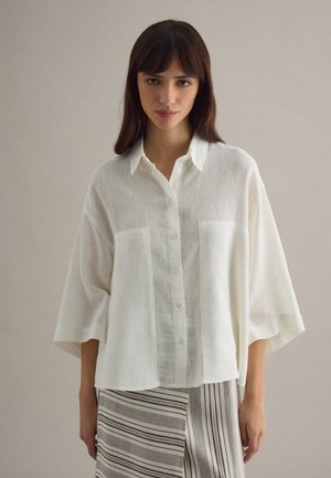 Joven mujer con una camisa blanca de botones holgada y mangas anchas, y una falda a rayas, posando frente a un fondo liso beige.