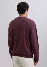 Sudadera burdeos con cuello redondo y puños acanalados, con una textura suave y un corte relajado, combinada con pantalones de color claro.