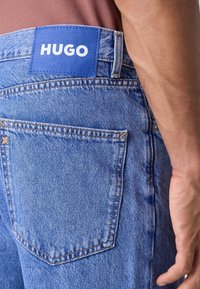 Jeans de mezclilla azul con un bolsillo trasero visible, que presenta una etiqueta azul con texto blanco "HUGO". La tela tiene una apariencia desgastada y costuras más claras.