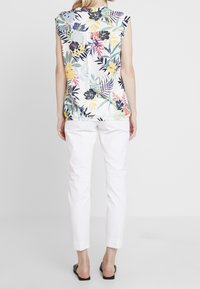 Top sin mangas floral con colores vibrantes sobre un fondo blanco, combinado con pantalones ajustados blancos y sandalias planas negras.