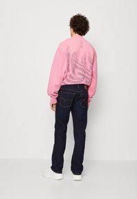 Sudadera rosa claro con gran texto estilizado en la parte posterior, combinada con jeans azul oscuro y zapatillas blancas. La vista trasera captura el diseño general del conjunto.