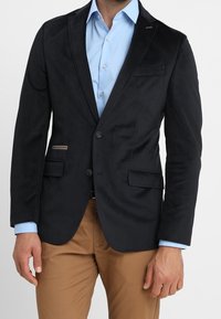 Svart blazer i texturerat tyg, med två framsidiga fickor, en enkel knappstängning och subtila detaljer på reversen. Ljusblå skjorta under.