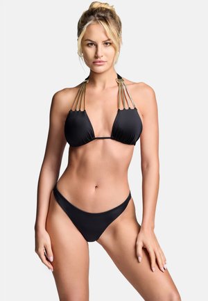 Kobieta o blond włosach, ubrana w czarne bikini z wieloma złotymi paskami na ramionach, stojąca na jednolitym białym tle.