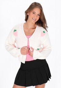 Witte gebreide cardigan met roze aardbeienpatronen, V-hals, en geribbelde manchetten, gedragen over een roze top en een zwarte geplooide rok.
