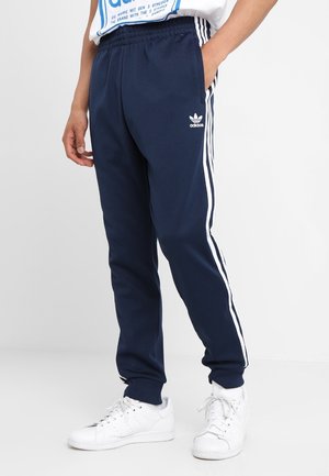Persoon draagt marineblauwe Adidas trainingsbroek met witte strepen en logo, gecombineerd met witte sneakers, staand tegen een effen achtergrond.