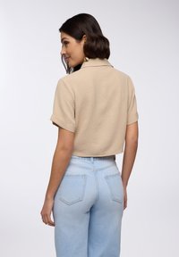 Camicia beige tagliata con superficie texturizzata e maniche corte; abbinata a jeans in denim azzurro chiaro a vita alta con taglio classico.