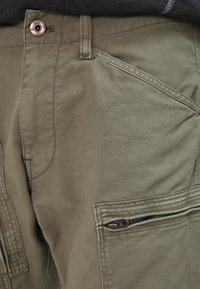 Olijfgroene cargo pants hebben een knoopsluiting, meerdere zakken, ritsdetails en een soepele katoenmix stof met een subtiele textuur.
