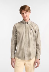 Giovane uomo che indossa una camicia button-up a righe verticali marroni e bianche e pantaloni beige, in piedi davanti a uno sfondo bianco semplice.