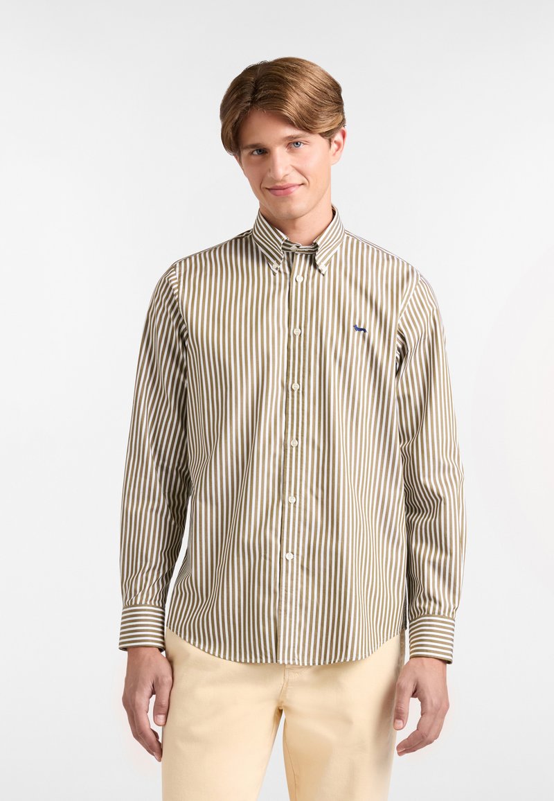 Giovane uomo che indossa una camicia button-up a righe verticali marroni e bianche e pantaloni beige, in piedi davanti a uno sfondo bianco semplice.