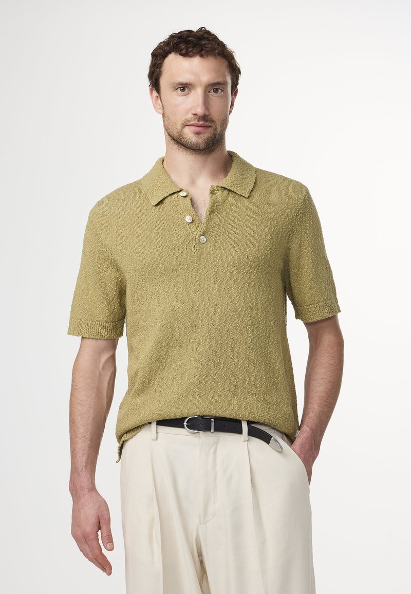 Korte mouwen polo shirt in gestructureerd olijfgroen katoen, met een kraag en drie knopen aan de voorkant, gecombineerd met crème broek en een zwarte riem.
