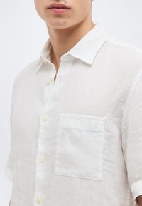Κοντινό πλάνο ενός ατόμου που φοράει λινό πουκάμισο με κουμπιά σε λευκό χρώμα, με γιακά και τσεπάκι στο στήθος, σε απλό φόντο.