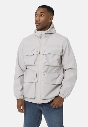 Mężczyzna w jasnoszarej kurtce z kapturem z czterema przednimi kieszeniami na klapy i niebieskich dżinsach, patrzący w bok na białym tle.