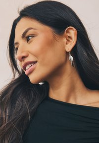 Silver dangling earrings with a wavy, elongated design. Smooth texture and polished finish. Worn by a person with long dark hair.