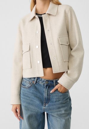 Vrouw met een beige cropped jasje met knopen en klepzakken over een zwarte cropped top en een high waist blauwe spijkerbroek, hand in de zak.