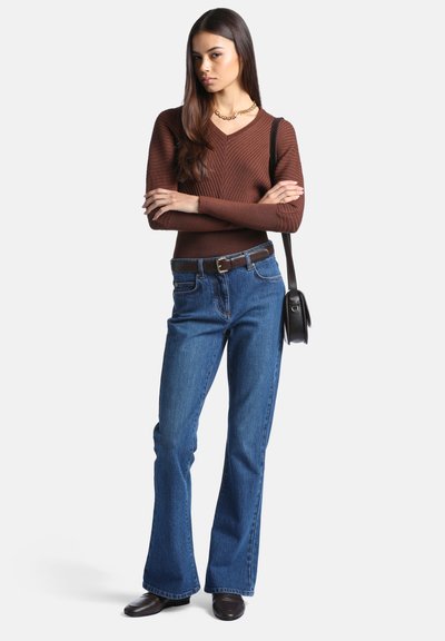 Femme debout, les bras croisés, portant un pull marron à col en V, un jean bleu, des chaussures noires, un collier en or et un sac à main noir
